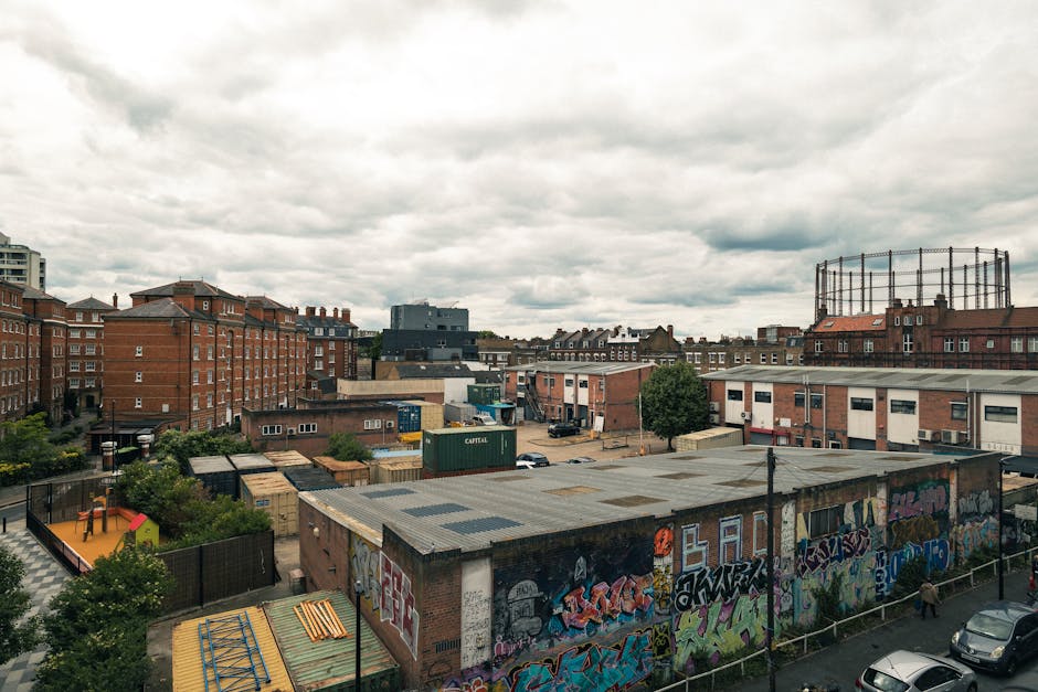 A view of an urban area taken from an elevated perspective under a cloudy sky, showing various buildings including brick apartment blocks, industrial warehouses, and a graffiti-covered structure with a flat roof. In the foreground, the flat roof of a low-rise building features faded graffiti art, and there are trees and small playground equipment visible near the fence. Behind, the area includes a parking lot with several parked cars, shipping containers, and industrial units, indicating an active commercial or storage zone. In the background, taller residential buildings, a large tank or silo, and additional industrial structures can be seen, with overcast weather casting diffuse lighting across the scene. This setting illustrates the environment in which [COMPANY_NAME] might perform house removals and furniture transport services, highlighting the urban relocation process and the logistics involved in moving within a cityscape.