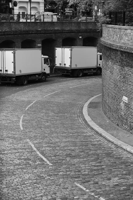 A black-and-white photograph depicts a narrow, curved brick-paved street with two medium-sized moving vans parked adjacent to a brick wall on the right side. The vans are positioned parallel to the curb, with their rear doors facing toward the street, suggesting an active loading or unloading process associated with house removals. The street is bordered by a curved brick wall on the right, and an open area with a bridge or overpass featuring multiple arches and a railing on the left. The overpass has trees and traffic signs visible on top, indicating an urban environment. The scene shows no people present, but the presence of the vans, packed inside and ready for transport, aligns with the process of home relocation and furniture transport. The overall setting emphasizes the logistical aspect of removals, with an emphasis on careful packaging and vehicle loading in a typical cityscape environment.