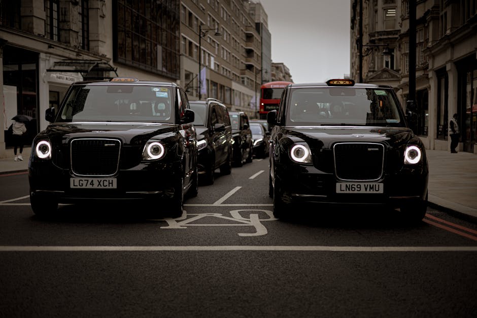 Two black London taxis with illuminated headlights are parked side by side on a city street in Hackney, with tall, historic buildings lining the road. Behind the taxis, there are several other vehicles, including a red double-decker bus, and pedestrians walking along the pavement on both sides of the street. The scene appears to be during late afternoon or early evening, with overcast grey skies. The area shows signs of urban activity typical of an active neighbourhood, with shopfronts and balconies visible on the buildings. This image captures a moment of typical city transport and street life, with the taxis possibly involved in home relocation or furniture transport services, as part of a house move or moving logistics in Hackney, coordinated by Removal Companies Hackney for residential moves and packing and loading processes.
