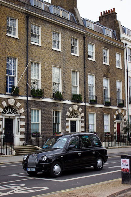 Two black London taxis with illuminated headlights are parked side by side on a city street in Hackney, with tall, historic buildings lining the road. Behind the taxis, there are several other vehicles, including a red double-decker bus, and pedestrians walking along the pavement on both sides of the street. The scene appears to be during late afternoon or early evening, with overcast grey skies. The area shows signs of urban activity typical of an active neighbourhood, with shopfronts and balconies visible on the buildings. This image captures a moment of typical city transport and street life, with the taxis possibly involved in home relocation or furniture transport services, as part of a house move or moving logistics in Hackney, coordinated by Removal Companies Hackney for residential moves and packing and loading processes.