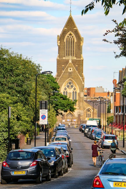 A narrow street in Dalston E8 lined with parked cars on both sides, including sedans and hatchbacks in various colours, with a woman walking a bicycle in the foreground. Behind the parked vehicles, there are a few pedestrians, some carrying moving boxes and bags, suggesting a home relocation process. The street is adjacent to residential buildings with brick facades and shopfronts, and the scene is set during daylight with partly cloudy skies. In the background, a prominent historic church with a tall, pointed stone steeple and arched windows is visible, surrounded by greenery from trees lining the street. Streetlights and traffic signs are also present along the pavement. This scene depicts a typical urban environment suitable for local removals and moving services by companies such as Removal Companies Hackney, capturing both the activity of loading and the setting for moving logistics in a residential neighbourhood.