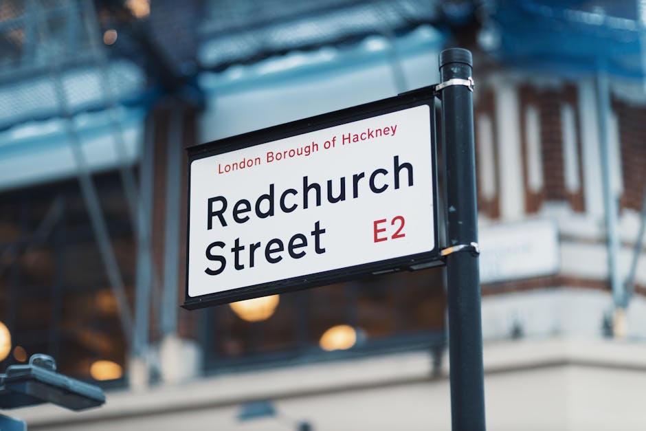 Close-up of a street sign reading 'Redchurch Street, E2' in London Borough of Hackney, with a white background, black text, and red postcode. The sign is mounted on a black metal pole, positioned on a busy street with buildings in the background featuring brickwork and large windows. Part of a scaffolding structure with blue netting is visible above the sign. The scene is well-lit with natural daylight, capturing a typical urban environment suited for house removals and furniture transport. The image is relevant for discussions on relocation logistics, parking tips, or street access concerns for removals in Hackney, as depicted by Removal Companies Hackney.