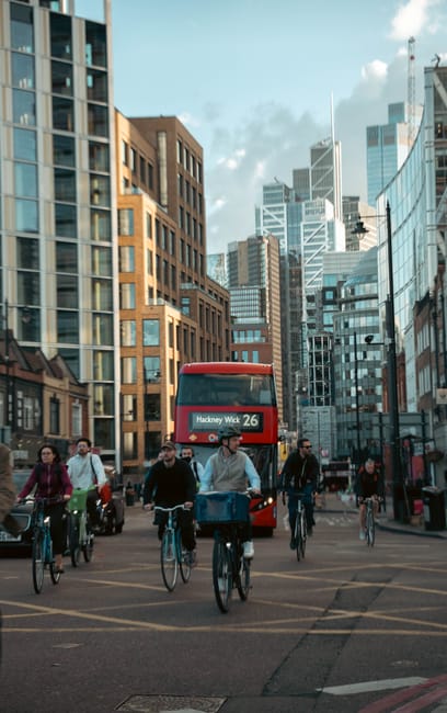 A red double-decker bus with the route number 26 and destination Hackney Wick is approaching a busy city intersection in Hackney, surrounded by tall modern office buildings and high-rise apartments. T