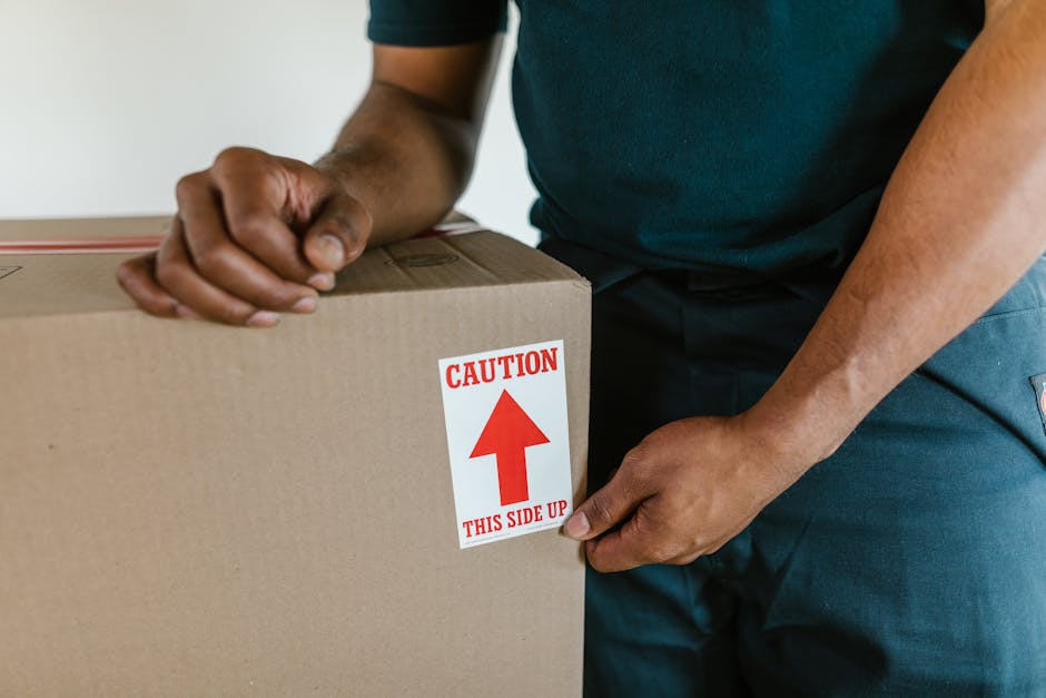 A close-up view of a person wearing a dark green shirt and dark trousers, handling a large cardboard moving box marked with a red and white caution label indicating 'This Side Up' and an upward arrow. The individual is holding the box securely from the side, with their hands positioned on the edges to ensure proper handling during the home relocation process. The scene appears to be inside a property, possibly in a corridor or near a doorway, with the focus on the careful alignment and lifting of the box. The background is plain and out of focus, emphasizing the handling of the packing box. This image illustrates furniture transport and packing and moving activities undertaken by Removal Companies Hackney as part of their same day removals service, highlighting the importance of proper labeling and cautious handling during loadings and unloadings in the relocation process.