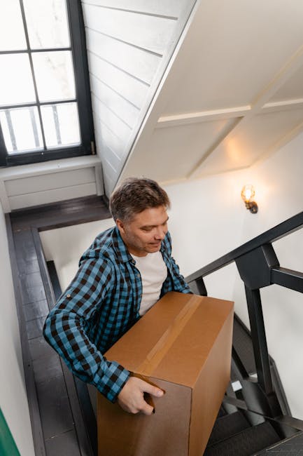 A man wearing a blue and black checkered shirt is carrying a large cardboard box with packing tape along its edges, navigating down a staircase inside a home. The staircase is dark wood with black metal railings, and the walls are painted white. Near the top of the stairs, a window with a black frame allows natural light to illuminate the area, complemented by a wall-mounted light fixture. The setting appears to be part of a home relocation process, with the man carefully managing the box during the loading or unloading of household items. This scene is captured indoors, reflecting the careful handling involved in house removals, especially when moving items through confined staircases. The environment is clean and well-lit, indicative of professional removals services such as those offered by Removal Companies Hackney, who specialise in detailed and safe furniture transport and packing during home moves.