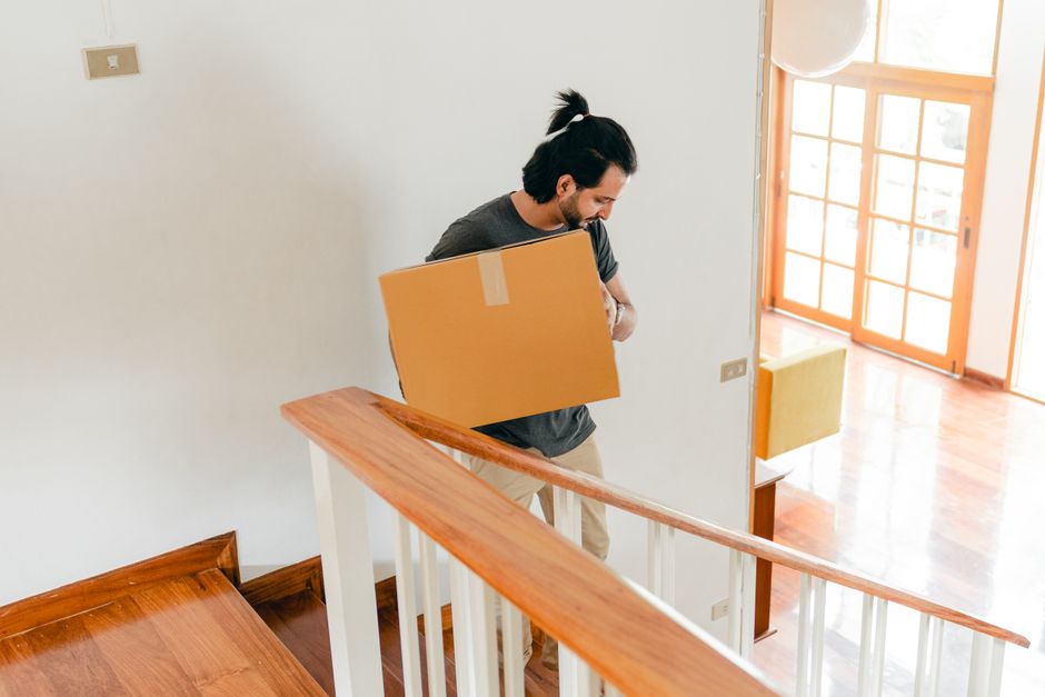 A man with dark hair tied back, wearing a dark grey t-shirt and beige trousers, is carrying a medium-sized cardboard box across a wooden staircase with white balustrades inside a residential property. The staircase is within a well-lit room, with natural light coming through large glass doors that lead to an outdoor space. The background shows a wooden floor, a yellow chair, and a white wall with a light switch. The image captures the process of a home relocation or furniture transport as part of a professional remova ls service, with the man carefully maneuvering the box during the loading or unloading phase in a house. The scene emphasizes careful handling and the indoor environment during a typical house move, supporting the context of packing, moving logistics, and furniture transport that Removal Companies Hackney provides within their removals service category for tight stair moves.