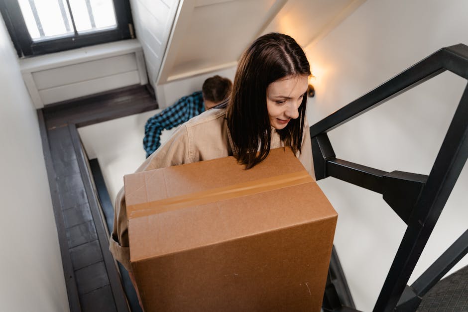 A man wearing a blue and black checkered shirt is carrying a large cardboard box with packing tape along its edges, navigating down a staircase inside a home. The staircase is dark wood with black metal railings, and the walls are painted white. Near the top of the stairs, a window with a black frame allows natural light to illuminate the area, complemented by a wall-mounted light fixture. The setting appears to be part of a home relocation process, with the man carefully managing the box during the loading or unloading of household items. This scene is captured indoors, reflecting the careful handling involved in house removals, especially when moving items through confined staircases. The environment is clean and well-lit, indicative of professional removals services such as those offered by Removal Companies Hackney, who specialise in detailed and safe furniture transport and packing during home moves.